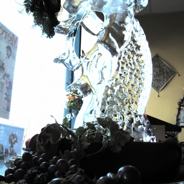 An ice sculpture of two figures stands behind a dark platter of grapes and other food items.